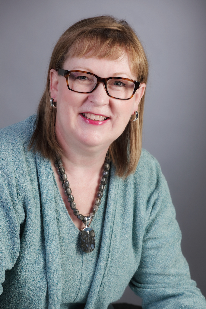 Head Shot of Helen Horton with short, straight, strawberry blond hair with glasses and blue-green blazer and warm smile.