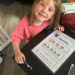 Gemma Bastian shows her letters. Gemma has straight blonde hair. She's wearing a salmon colored shirt. She's got a pen in her and and a tracing card with her name on it.