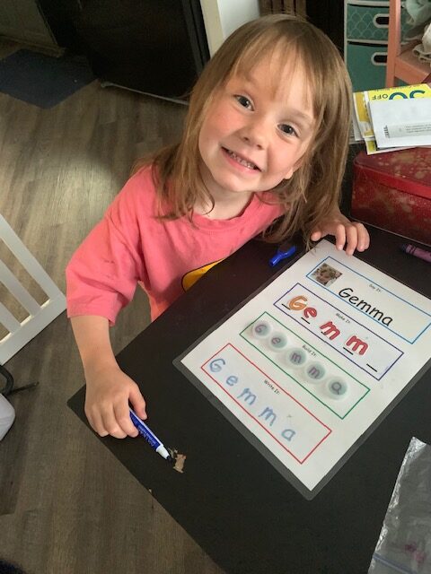 Gemma Bastian shows her letters. Gemma has straight blonde hair. She's wearing a salmon colored shirt. She's got a pen in her and and a tracing card with her name on it.