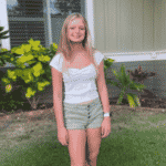Taylor Sandon stands on a lawn. She has straight blonde hair and wears a white top and khaki shorts in front of a flower bush.
