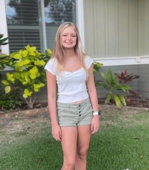 Taylor Sandon stands on a lawn. She has straight blonde hair and wears a white top and khaki shorts in front of a flower bush.