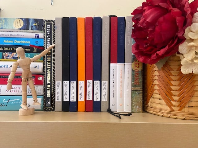 Several colors of journals on the shelf. A wooden person pointing to the multi-colored journals.