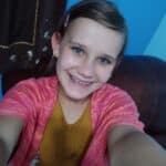 Addison Call is in a mustard colored shirt with pink cardigan. Her hair is back with a pink barret. The background is different shades of blue. She has braces and earrings. Blonde hair and blue eyes. She's holding the camera for a selfie.