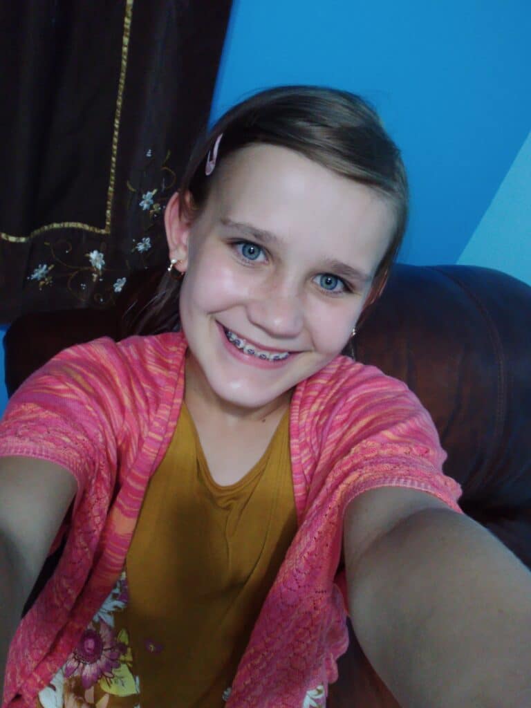 Addison Call is in a mustard colored shirt with pink cardigan. Her hair is back with a pink barret. The background is different shades of blue. She has braces and earrings. Blonde hair and blue eyes. She's holding the camera for a selfie.