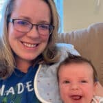 Katy Meeks and Baby Luke. Katy's straight blonde hair droops against her shoulders and wearing a blue and green hoodie. She's wearing glasses and smiling. Her baby boy Luke gives a big open smiles to the side in her arms. A burping bib just above him. His hair sticks up.