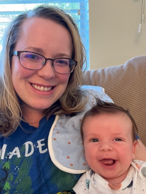 Katy Meeks and Baby Luke. Katy's straight blonde hair droops against her shoulders and wearing a blue and green hoodie. She's wearing glasses and smiling. Her baby boy Luke gives a big open smiles to the side in her arms. A burping bib just above him. His hair sticks up.
