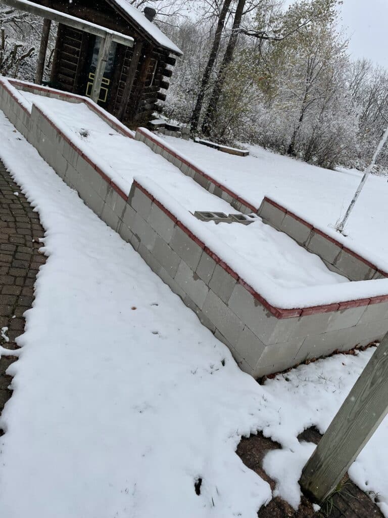 Snowy backyard. Bricks laid out in a terrace on the gentle slope of the yard covered with a half inch of snow. Red bricks rim the top. In the background there is a shed with dark brown logs. Behind it is a forrest.