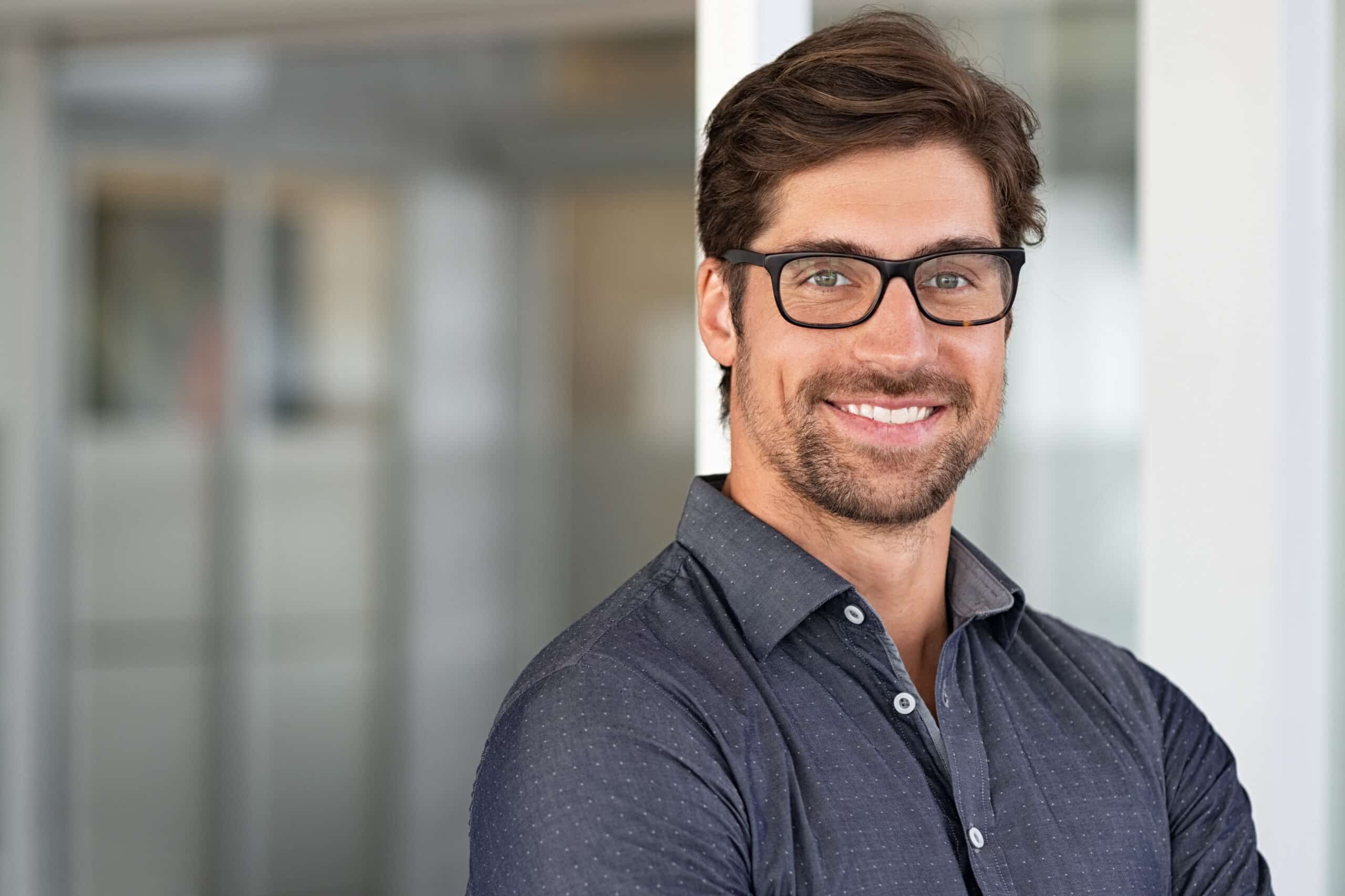 Portrait of handsome businessman standing in modern conference room. Happy young business man in shirt looking at camera with copy space. Portrait of successful smiling guy in office wearing eyeglasses.